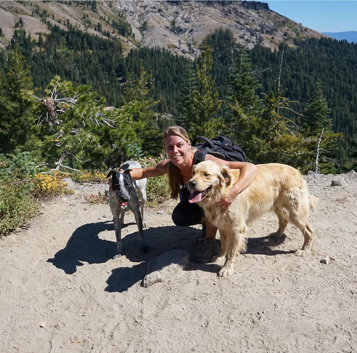 Woman crouched on a mountain trail with two dogs on leash – Hiker and pet sitter poses with two dogs in a scenic mountain setting, showing outdoor bonding. Southern California Pet Sitting 