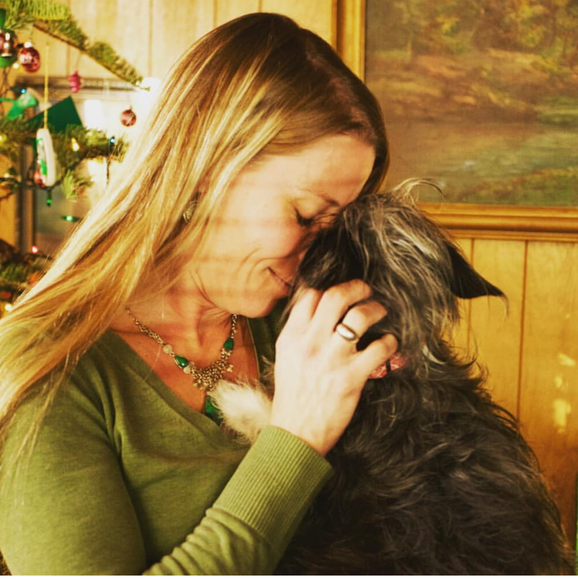 Woman with blonde hair and green sweater cuddles with Small brown dog with wiry hair.  Southern California Pet Sitting 