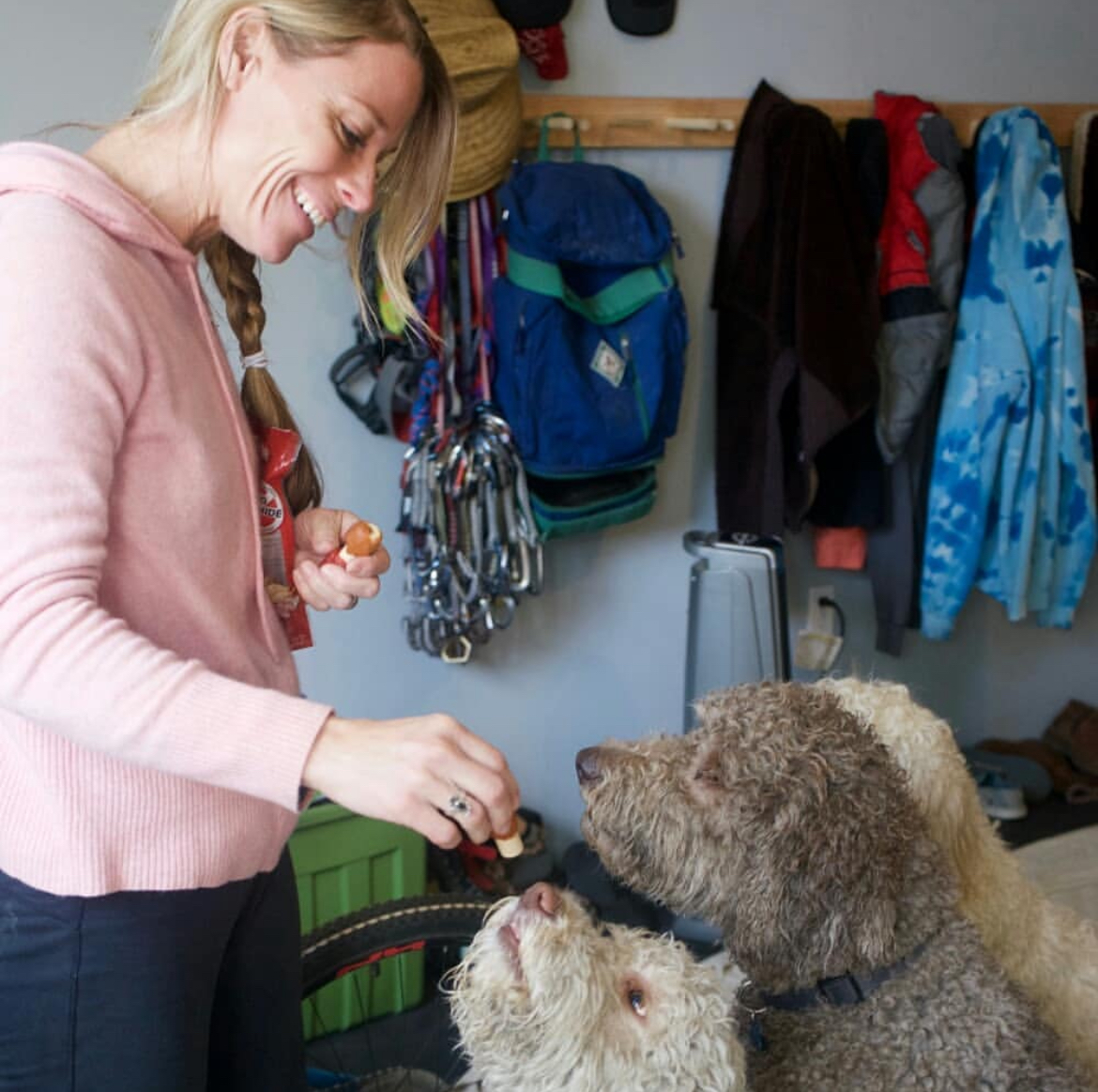 Blonde haired woman with a braid is feeding treats to three different dogs in a mudroom with gear in the back. Southern California Pet Sitting  