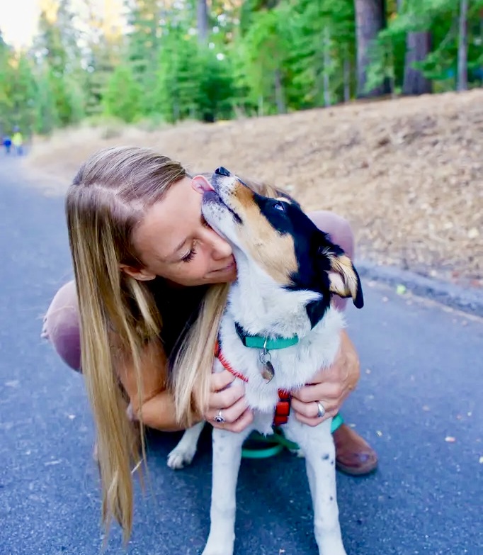 Woman crouched down kissing a mixed-breed dog on a wooded trail — tender bond between pet sitter and dog during nature walk. about Southern California Pet Sitting