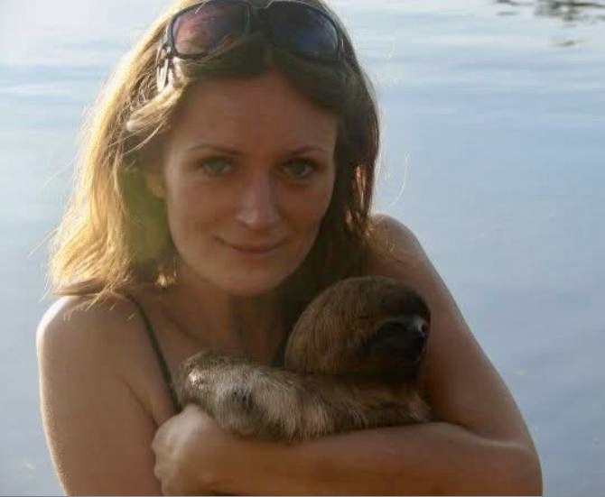 Woman holding a small brown sloth lovingly near the water’s edge at sunset. Southern California Pet Sitting 