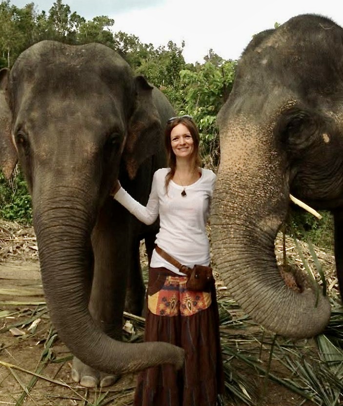 Woman standing calmly between two elephants, gently touching them in a forest setting..  exotic animal care, intuitive elephant connection, animal communication, global pet sitting experience, soul-level animal trust. about Southern California Pet Sitting