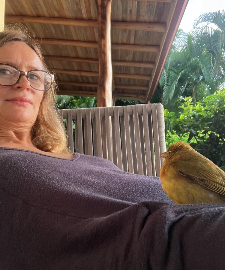 Woman with brown hair and glasses lays back with a small bird on her leg. Southern California Pet Sitting 